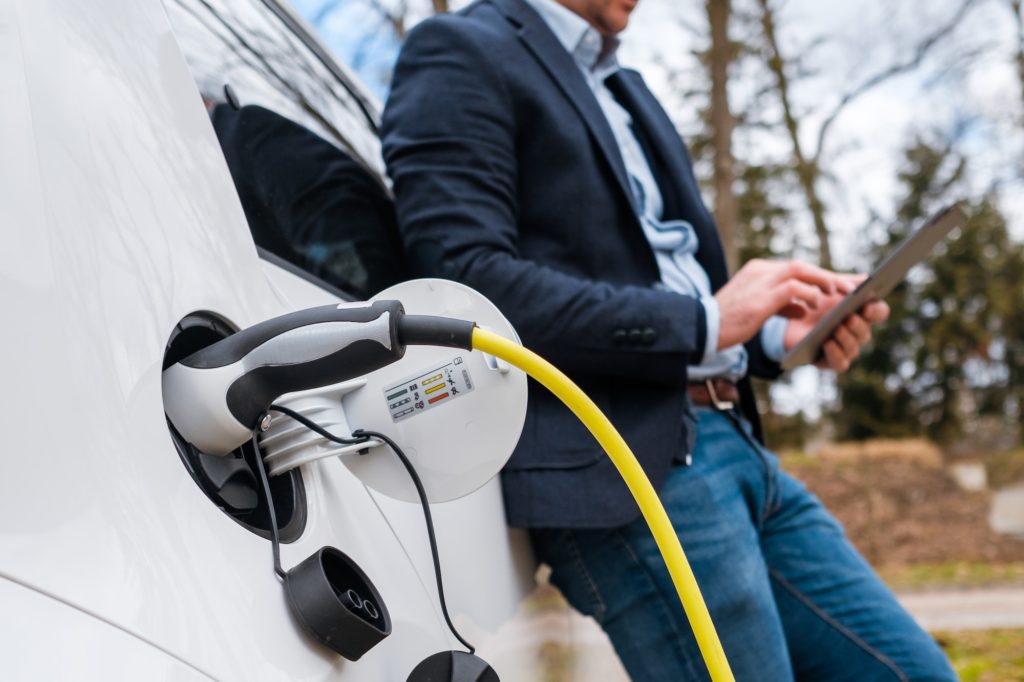 Business man standing near charging electric car or EV car and using tablet in the street