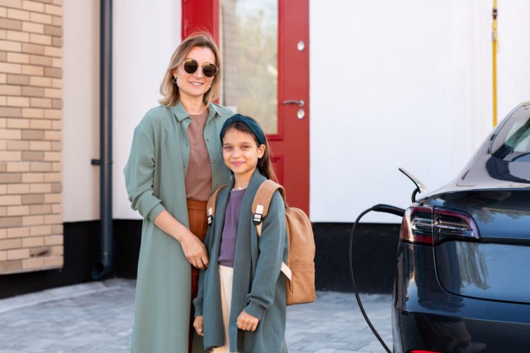 Happy young female and little schoolgirl standing by electric car